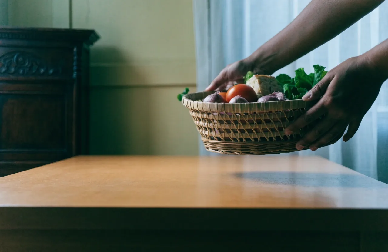 Hands moving a basket of market vegetables and tempeh across a wooden table